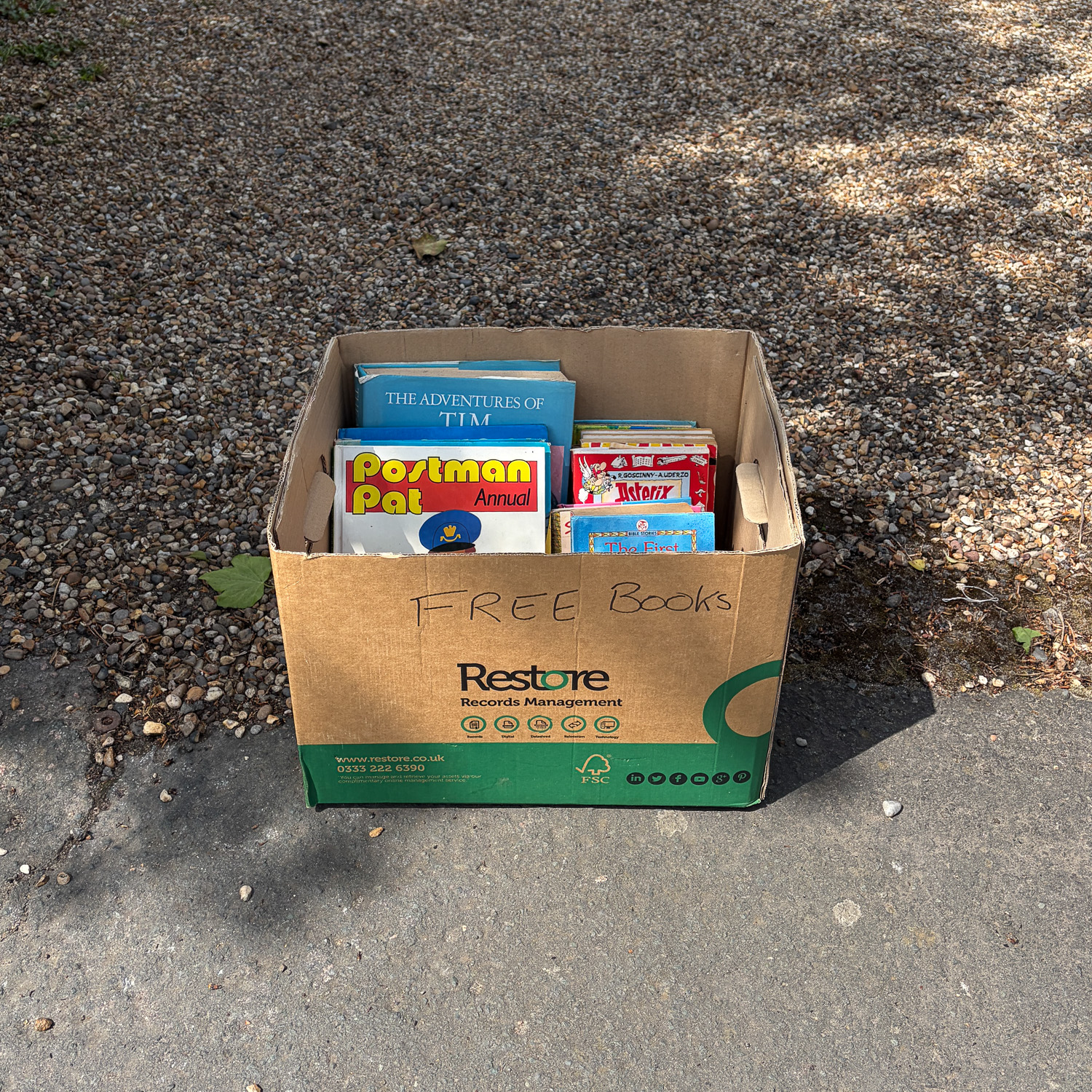 Photograph of a cardboard box with 'FREE BOOKS' handwritten on the front, with books including Postman Pat, Asterix and The Adventures of Tim'. There is a gravel area behind and pavement in front.