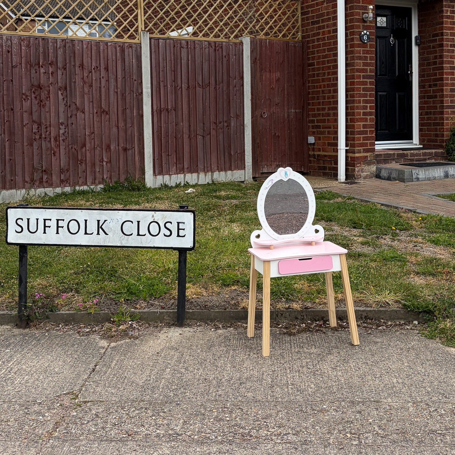 Photograph of a child's vanity table with mirror and pink drawer at the edge of a pavement, next to a sign that says 'SUFFOLK CLOSE'. Behind is a house with fence and small lawn and in the foreground is a concrete path.