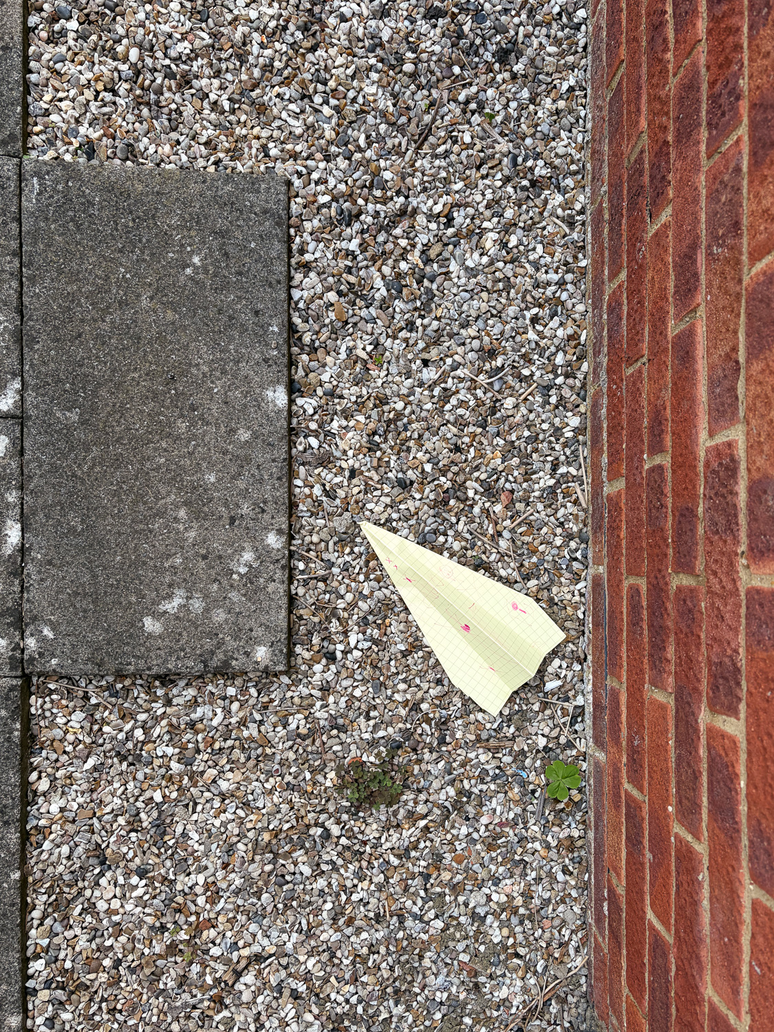 Photograph looking down at a gravel area with a paving slab to the left and brick wall to the right. In the centre is a paper airplane made from yellow graph paper with children's drawing on it.