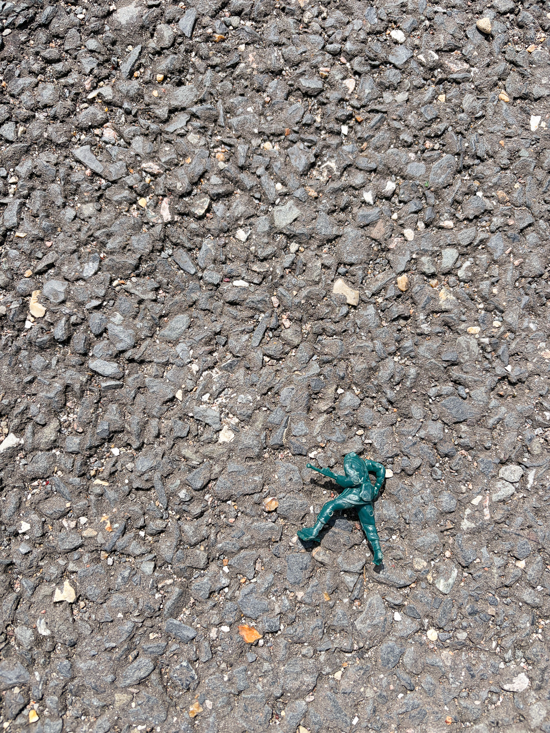 Photograph looking down at an asphalt path, with a scuffed and headless green plastic toy soldier just below the centre of the frame.