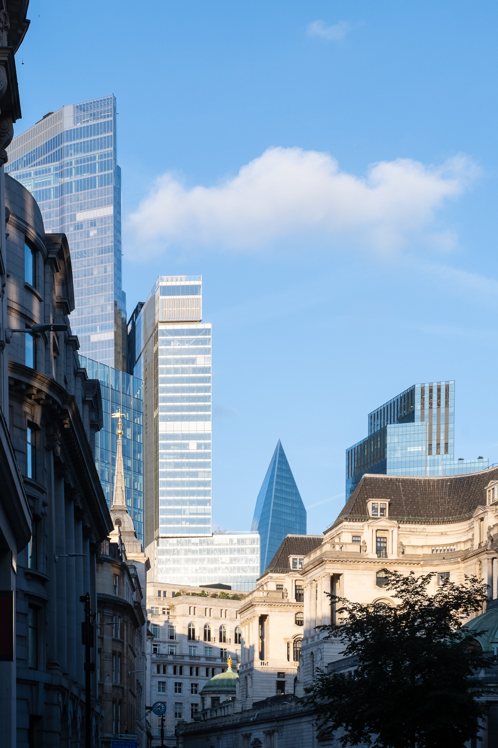 Photograph of a London skyline, with high-rise glass buildings in the background, with older buildings in the foreground. The sun is hitting the tops of the far buildings and there is shadow in the lower portion of the frame. Blue sky with a fluffy clouds is above and to the right.