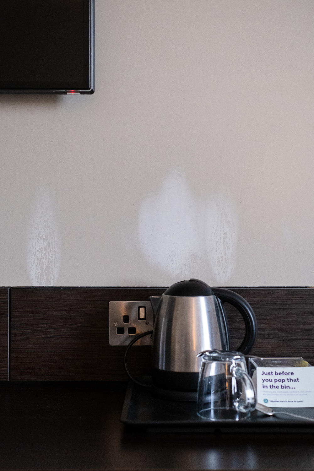 Photograph of a metal kettle on a tray on a desk with wooden panel with plug socket behind. Also on the tray is a glass mug and a sign that says 'Just before you pop that in the bin...'. On the beige wall behind there are 3 white areas where steam from the kettle has stained the wall. At the top left of the frame part of a TV screen can be seen.