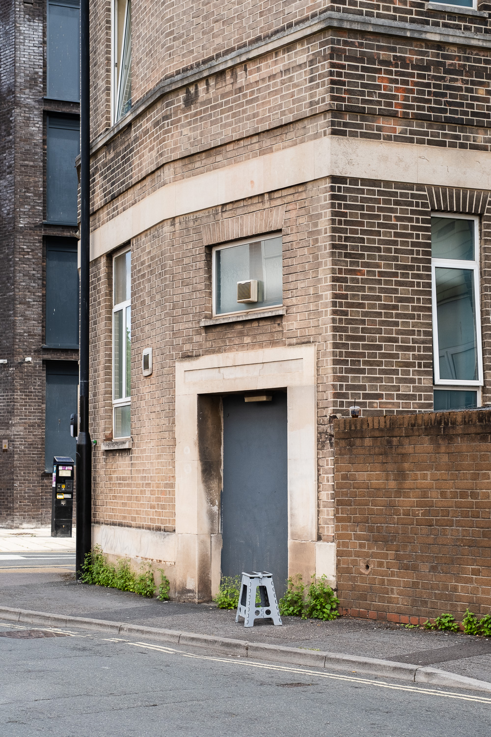 Photograph of a grey plastic foldable stool at the side of a road, on a pavement. Behind is a building with doorway that is boarded with a dark grey panel. The brick building is on a corner.