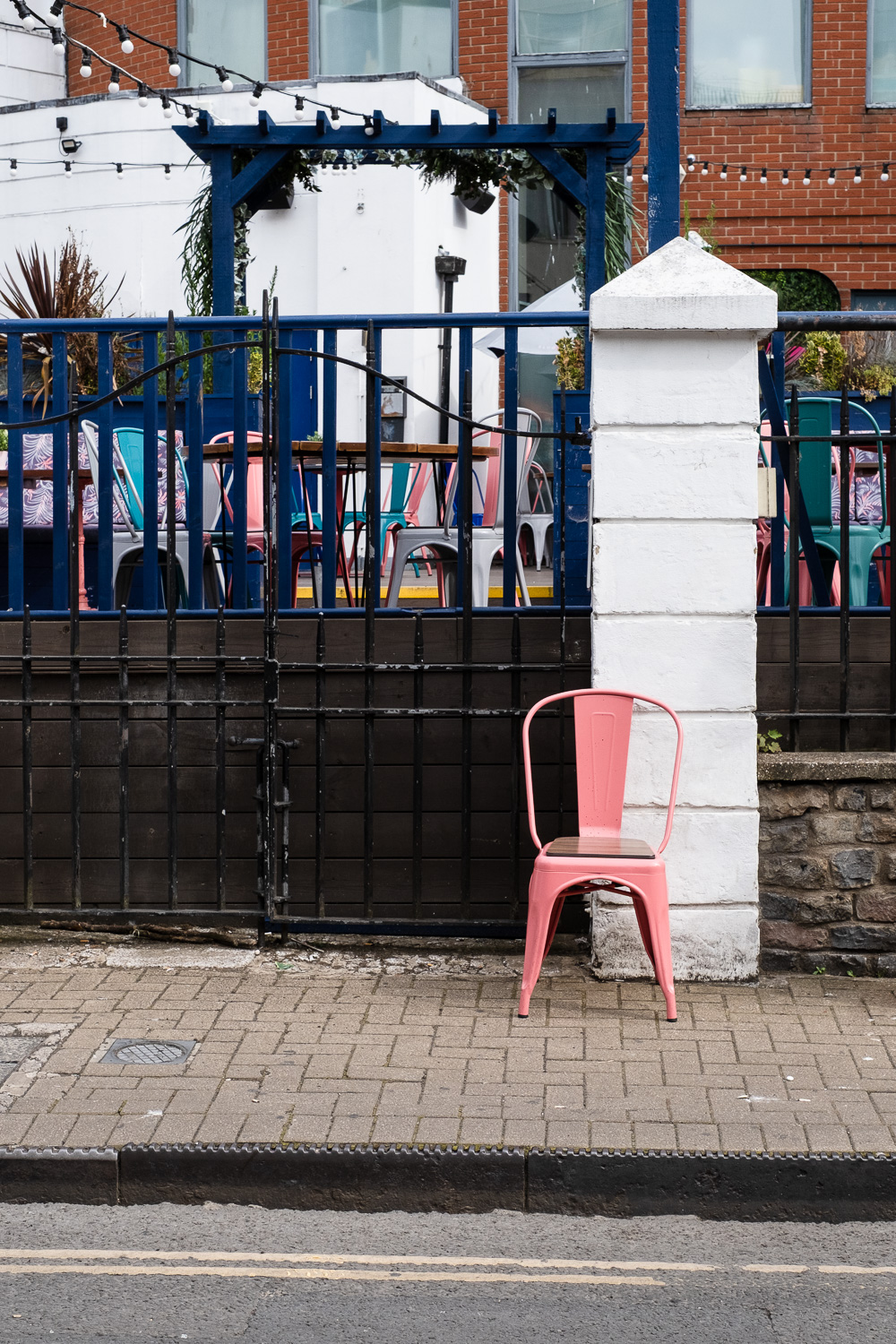 Photograph of a pink metal chair at the side of a road, on a paved pavement. Behind is a white pillar and metal railings with a seating area, blue pergola and festoon lights above and behind. In the foreground there is a metal kerb and double yellow lines on the road.