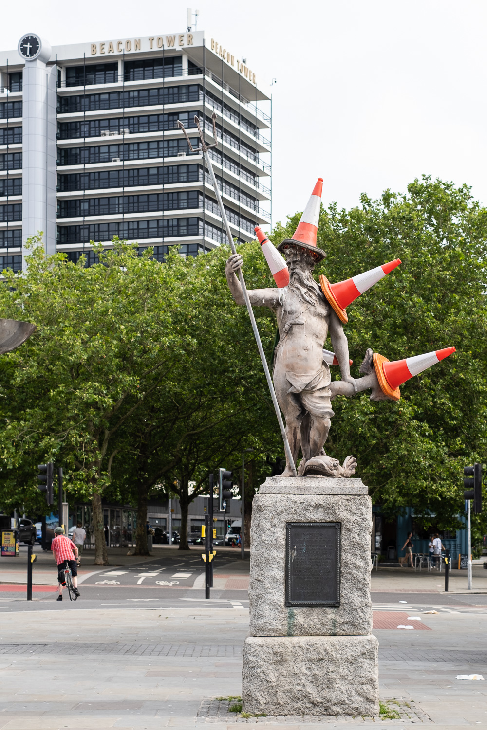 Photograph of the statue of Neptune, in Bristol, with orange and white plastic traffic cones placed on his head and other areas, 4 clearly visible, one behind. Behind the statue is a road with a cyclist on it, leafy trees and a high building with a clock and 'BEACON TOWER' at the top.