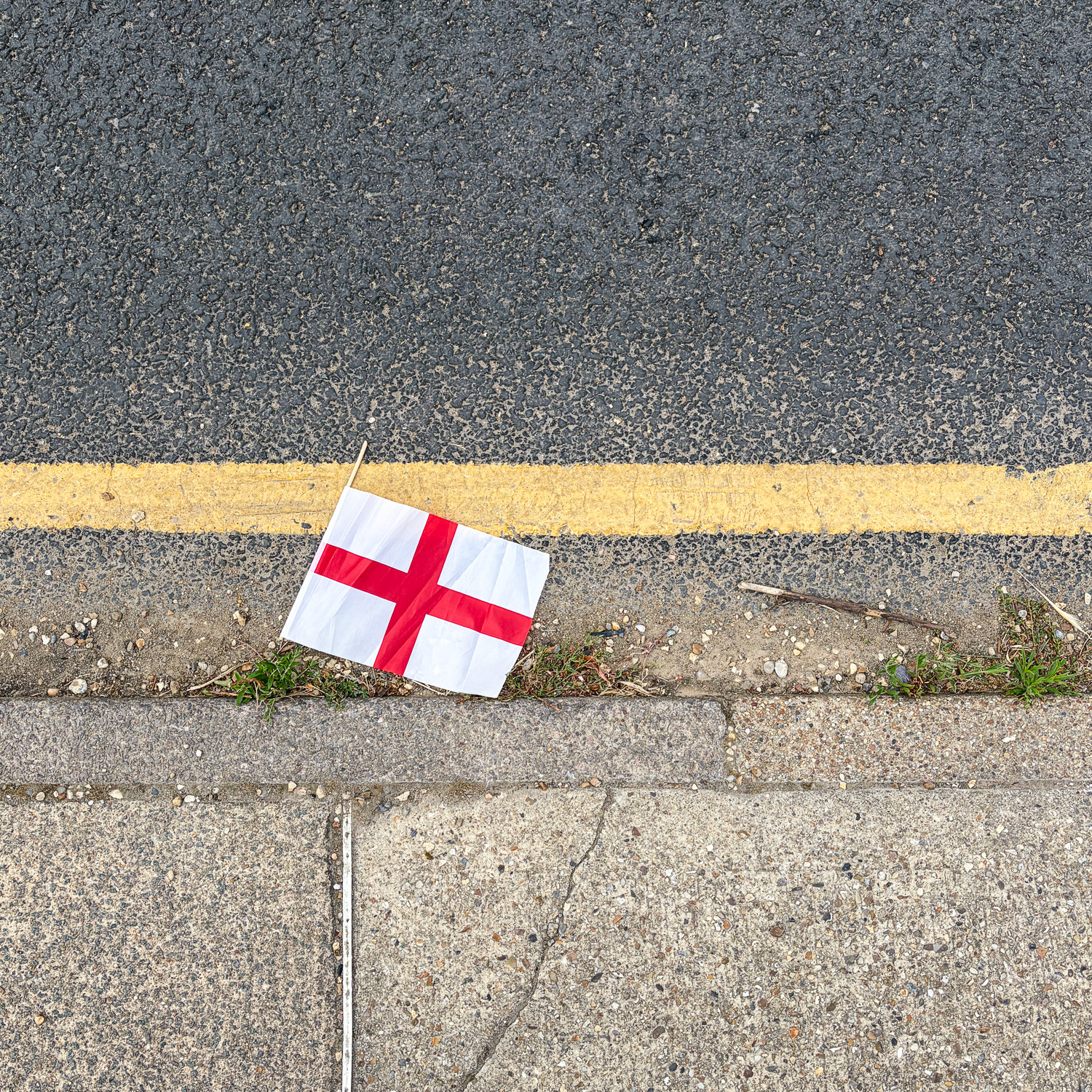 Photograph looking down at a gutter at the side of the road, with a small plastic St George's flag (red cross on white background) in the road. A single yellow line is painted on the road and a pavement with curb is at the bottom of the frame.