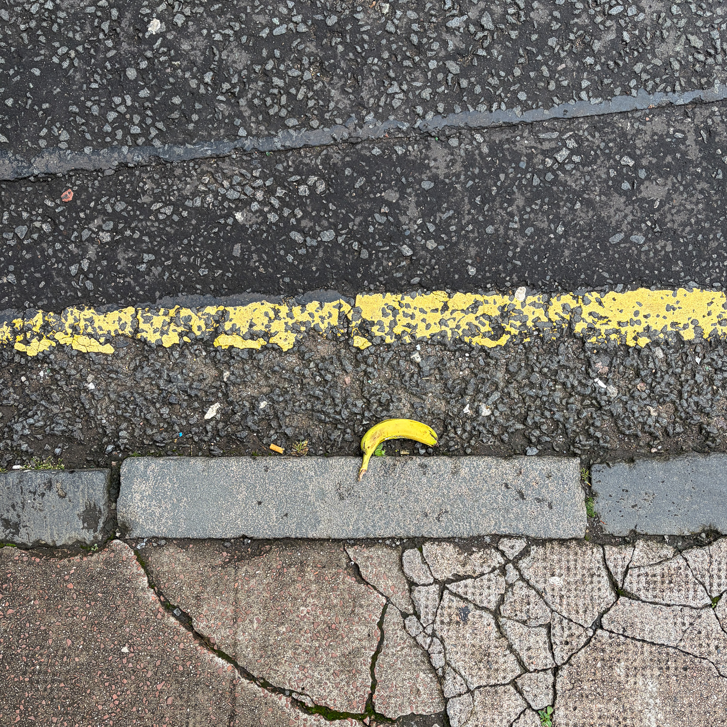 Photograph looking down at a gutter at the side of the road, with a banana against the curb, a single yellow line above and broken paving at the bottom of the frame.