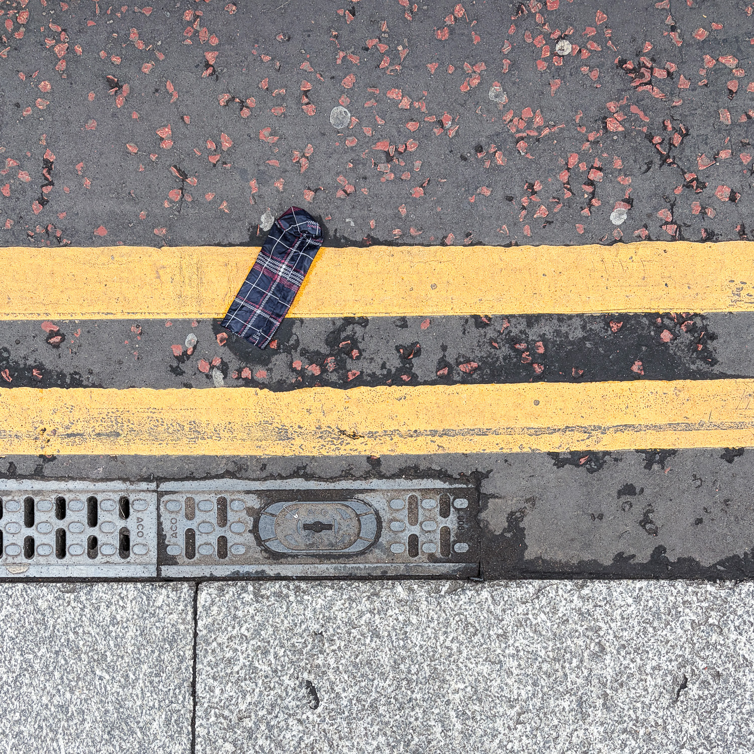 Photograph looking down at a gutter at the side of the road, with a wet tartan umbrella cover on 2 double yellow lines, with grate and paving slabs at the bottom of the frame.