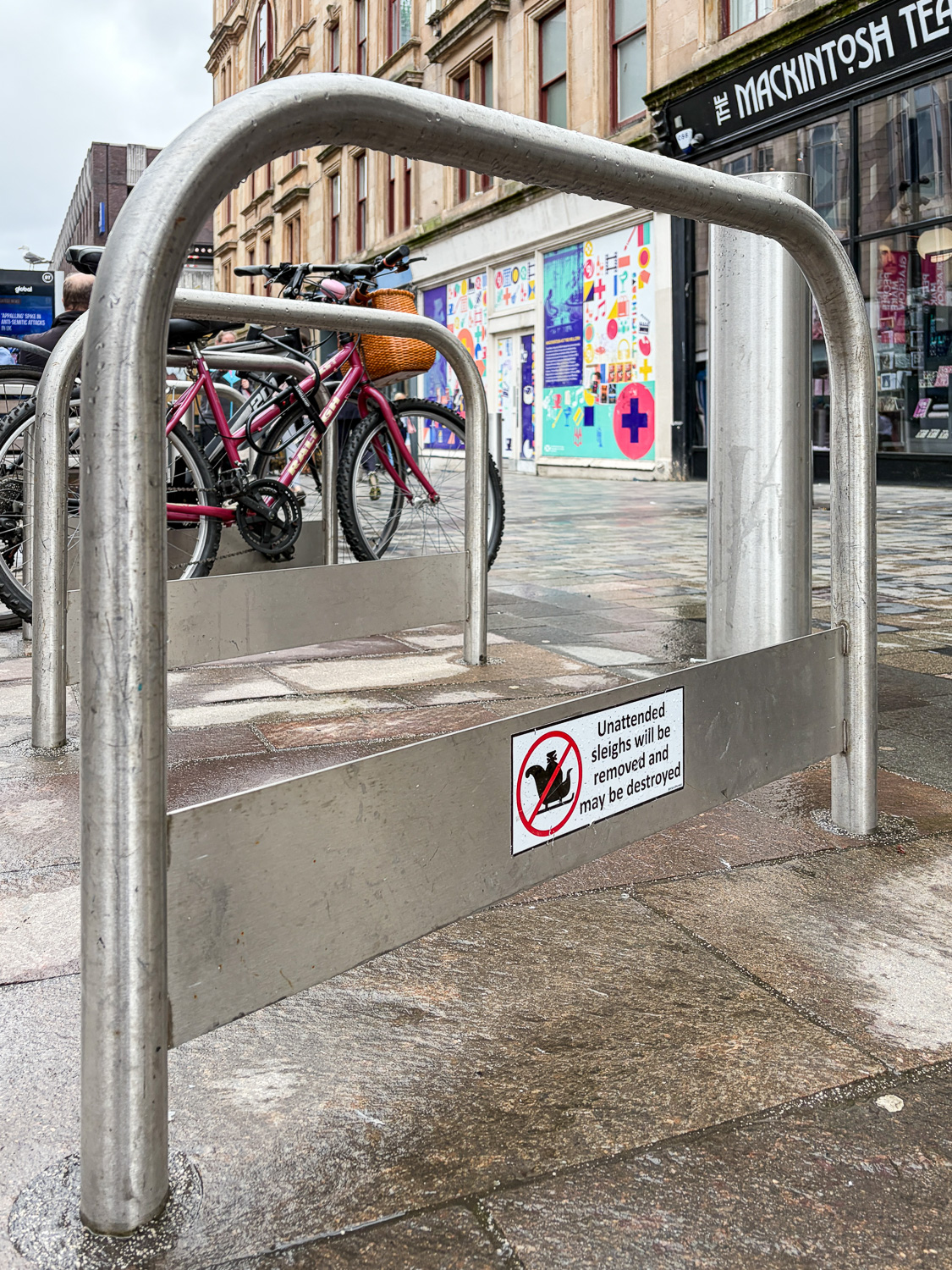 Photograph of a bike rail with a sticker on it with a logo of a sleigh with a red circle and cross through it, with 'Unattended sleighs will be removed and may be destroyed' to the right. Behind is a shopping street and parked bikes.