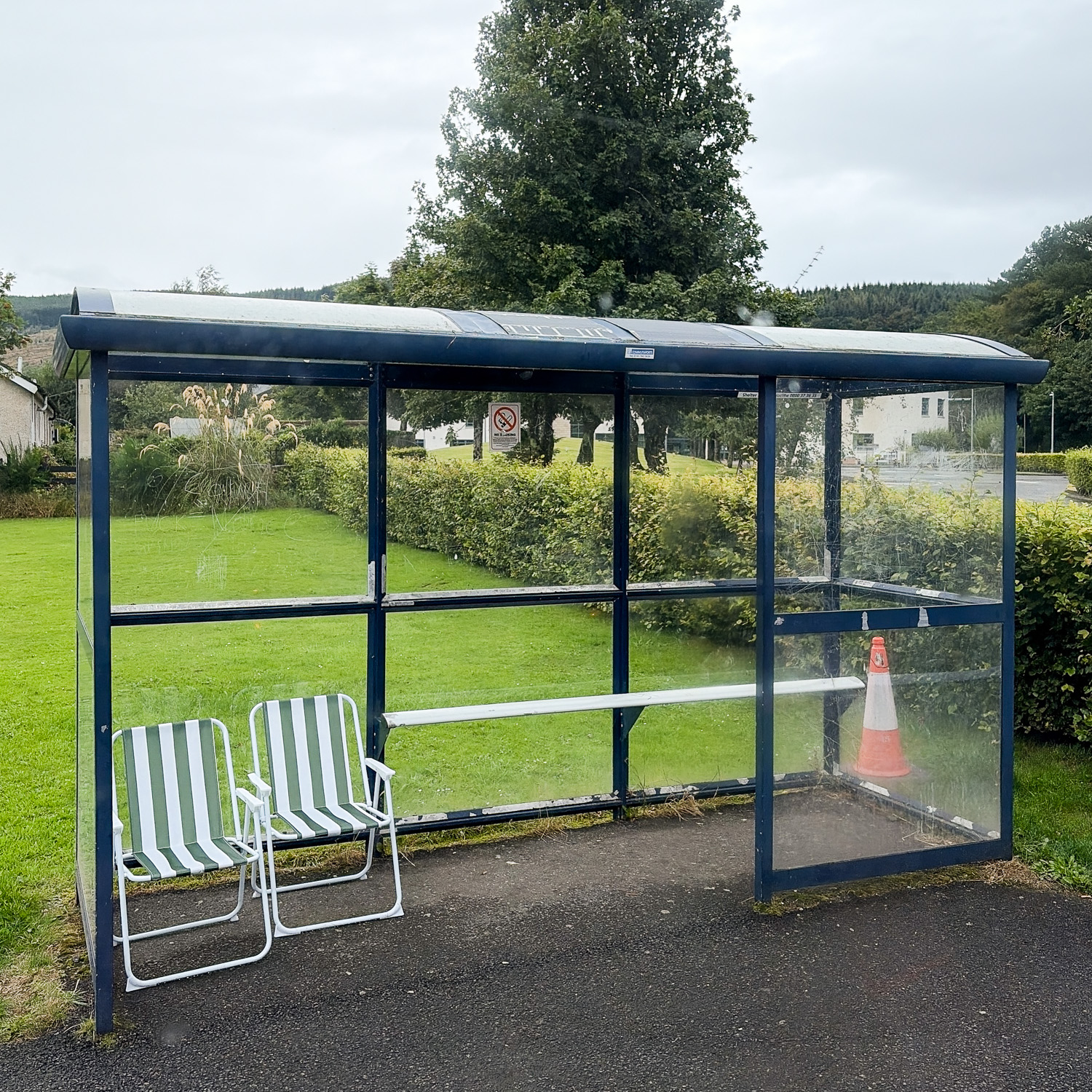 Photograph of a covered bus stop with two striped folding chairs inside to the left and a traffic cone to the right. There is a tarmac floor underneath and in front and lawn, hedge and trees behind.