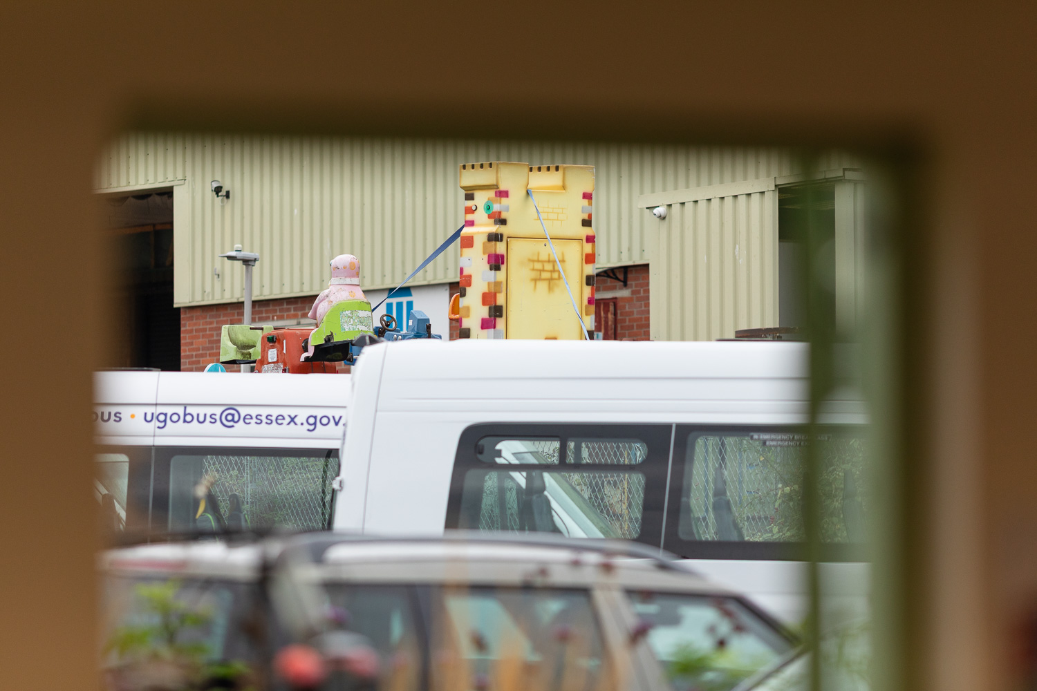 Photograph looking out through the top of a doorway, with the back of what appears to be a large plastic Mr Blobby ride up high, in front of a warehouse, with white vans and vehicles out of focus in front.