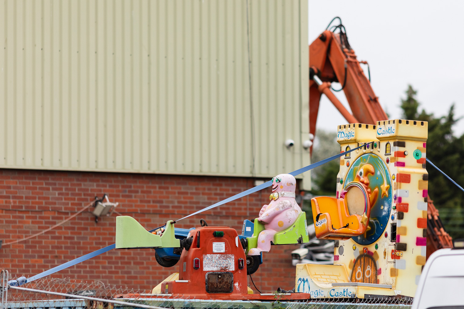 Photograph of a plastic Mr Blobby dee-saw ride with a another ride that has 'Magic Castle' written at the top and bottom behind it, in front of a warehouse, with part of an orange digger in the background behind.