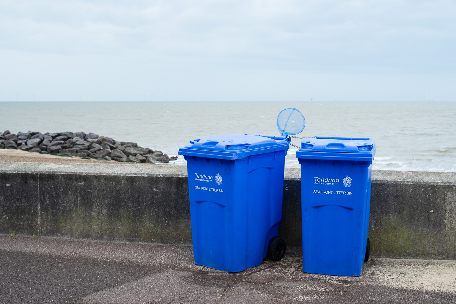 Photograph of two large blue wheelie bins with 'Tendring District council, SEAFRONT LITTER BIN' in blue writing, and a blue child's fishing net sticking out the top of the one on the left. Behind is a sea wall, pile of large rocks and the sea and sky in the distance.