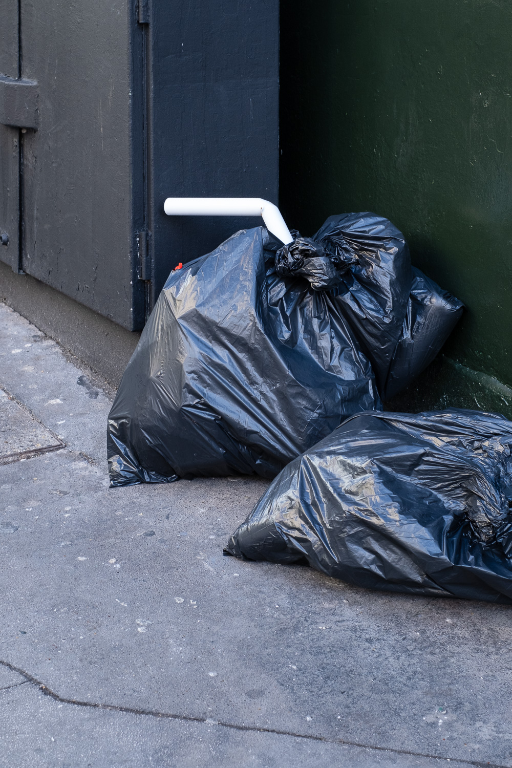 Photograph of a bent white plastic pipe sticking out of a black bin bag on a pavement, resembling a drinking straw, next to another bin bag.