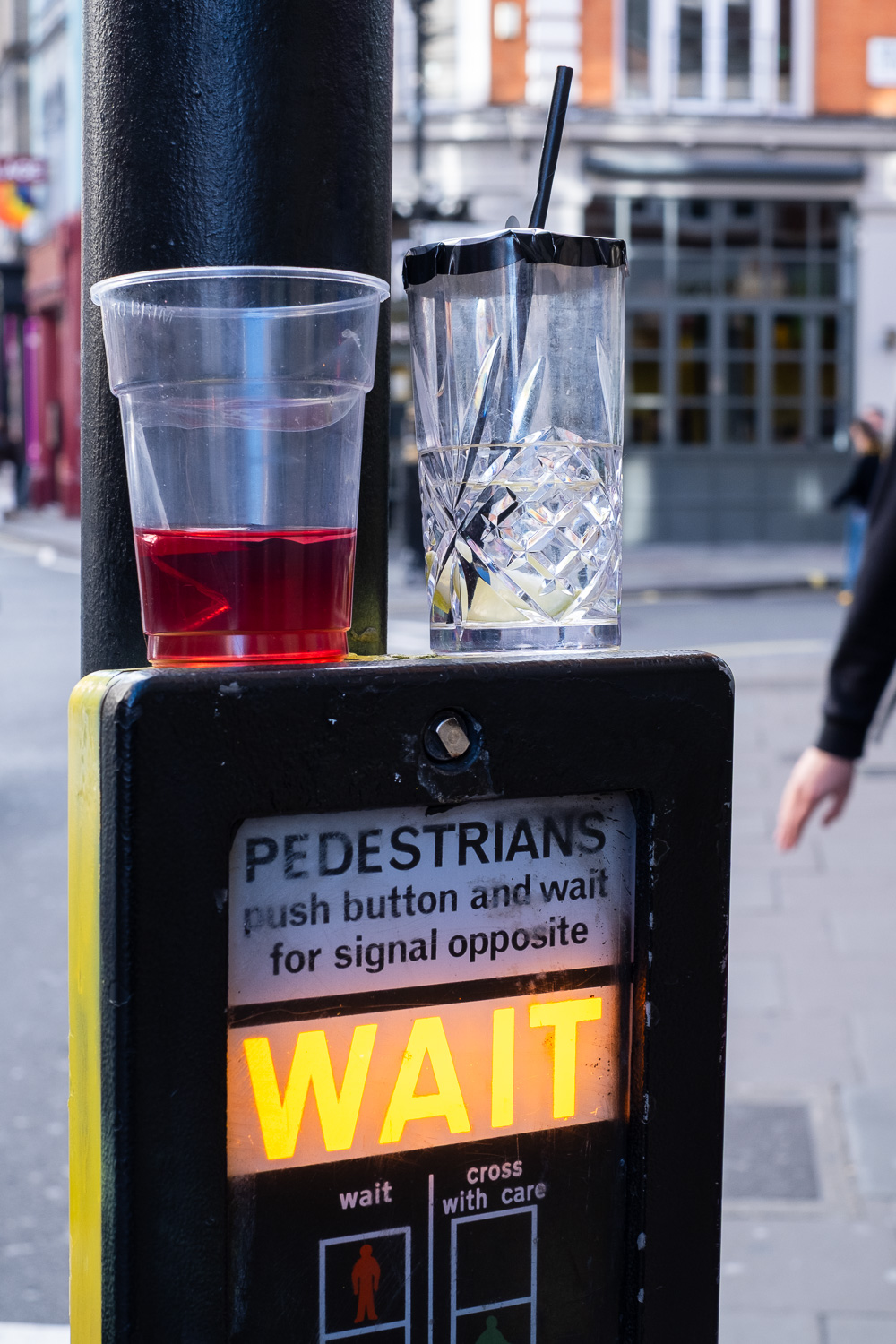 Photograph of two abandoned drinks on top of a 'PEDESTRIANS, push button and wait for signal opposite, WAIT' box. To the left is a clear plastic cup with a red liquid at the bottom and to the right is a cut glass glass with a clear liquid, black straw and black top to cover the top of the glass.