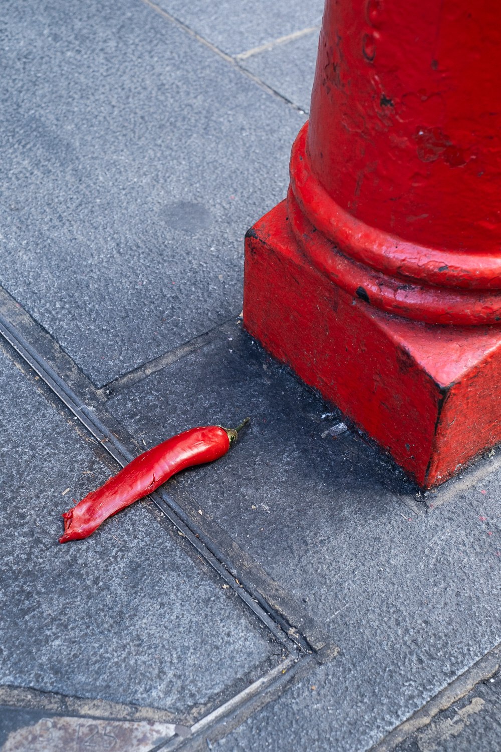 Photograph of a long, glossy red chilli with a squashed end, on the pavement next to the base of a bright red pillar to the right.