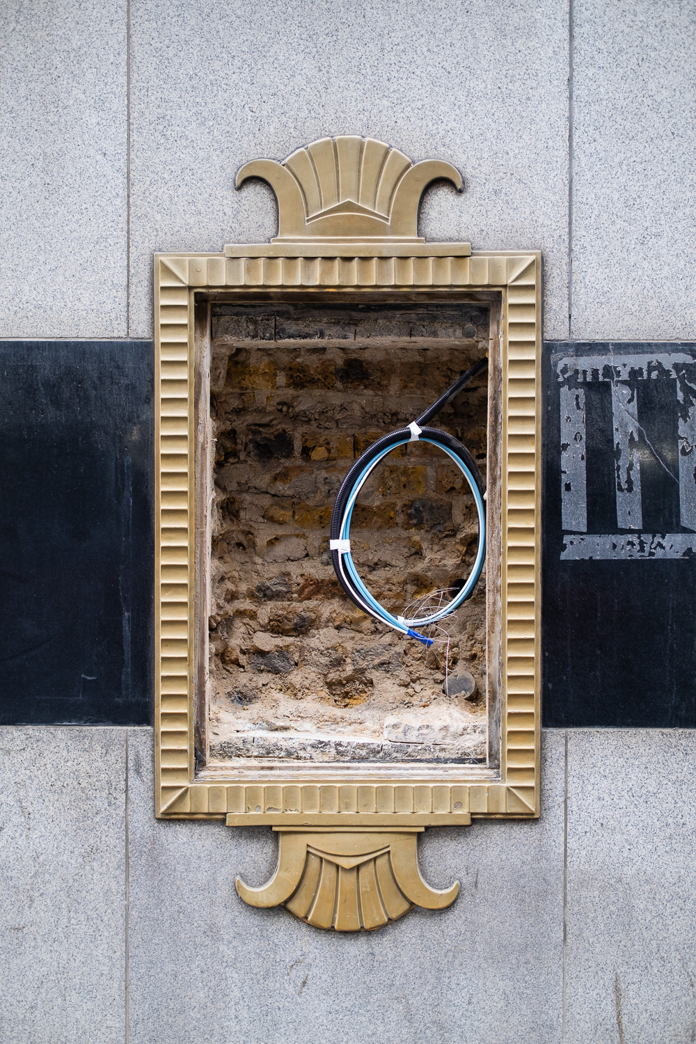 Photograph of an ornate gold frame on a tiled wall, with exposed brick and a coil of cables inside.